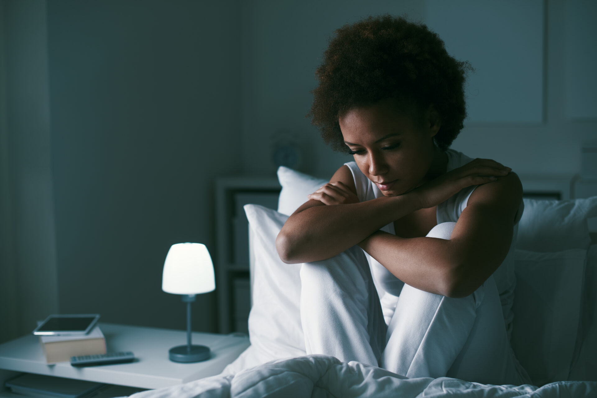 Woman sitting on bed at night, looking down and appearing thoughtful or concerned.